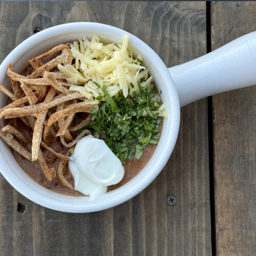 White bowl on a wood background filled with Chicken Tortilla Soup garnished with tortilla strips, cheese, cilantro, and sour cream.