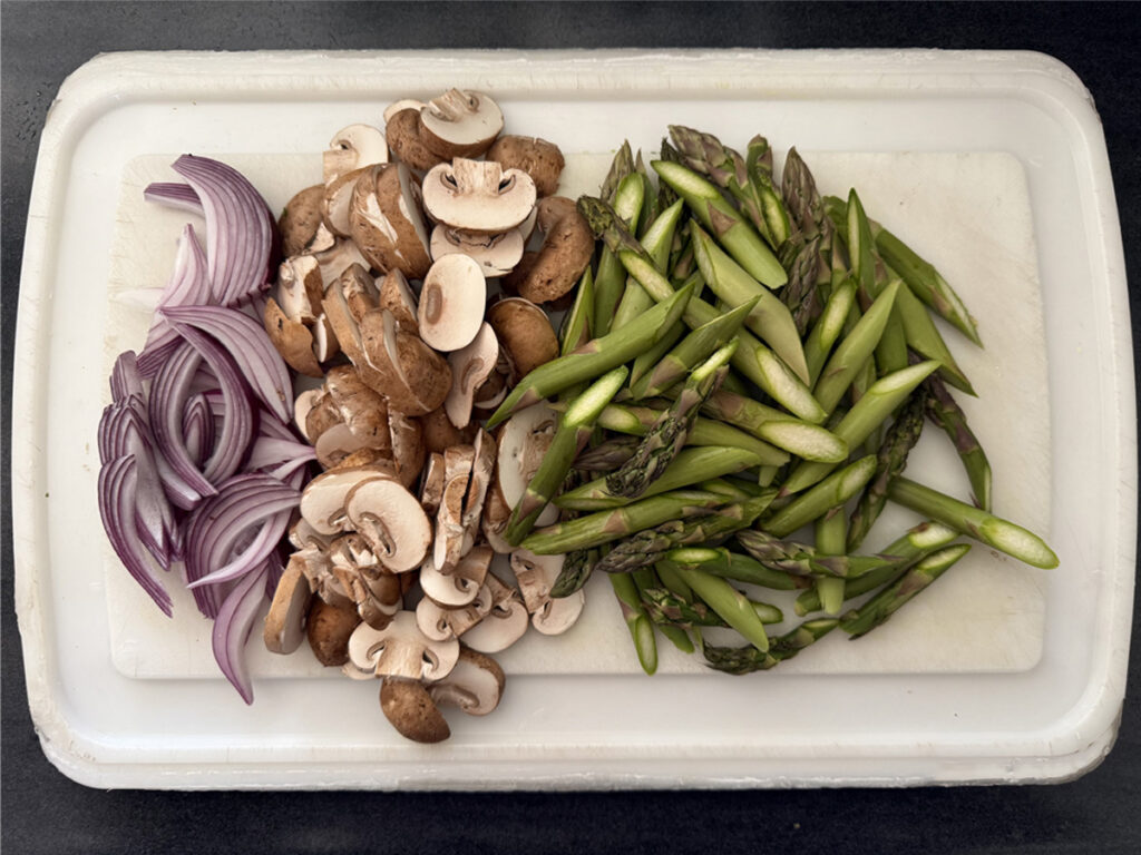 Asparagus, cremini mushrooms, and red onions sliced on cutting board.