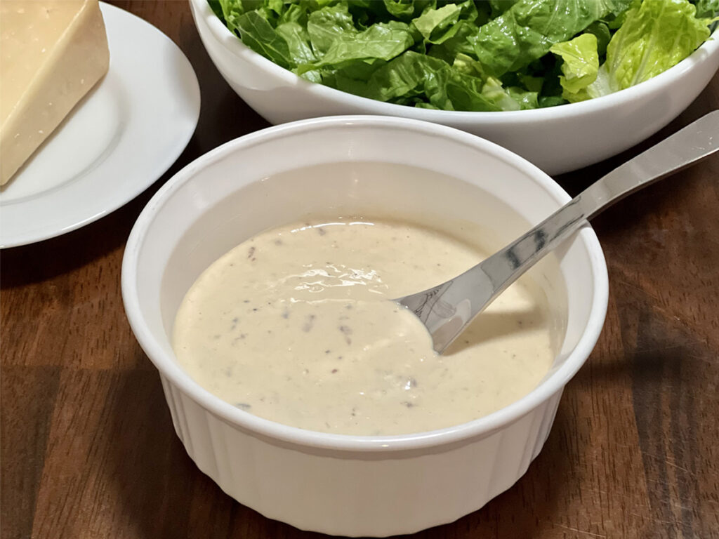 Caesar Salad Dressing (Outback Copycat) in a small white bowl with a glimps of Romaine and Parmesan cheese in the background.