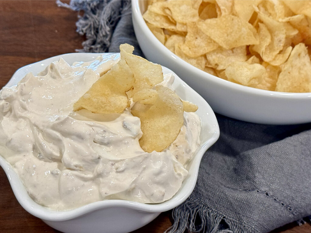 French Onion Dip with Caremlized Onions in a small white bowl with a few chips in the bowl plus more chips in the background.