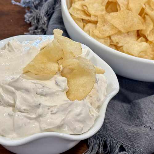 French Onion Dip with Caremlized Onions in a small white bowl with a few chips in the bowl plus more chips in the background.