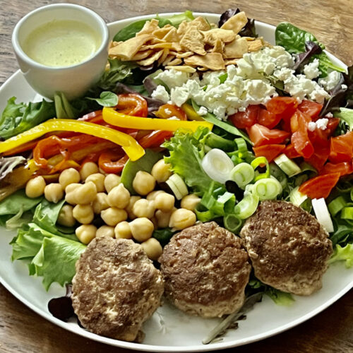 Colorful healthy mediterranean salad with lettuce, chickpeas, scallions, tomatoes, red and yellow pepper slices, feta cheese crumbles, crushed pita chips, three Greek meatballs and a ramekin of dressing.