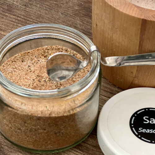 Easy Homemade Seasoned Salt in a clear jar with a small spoon on the edge and a white lid on the wood counter marked with a sticker Salt Seasoned.