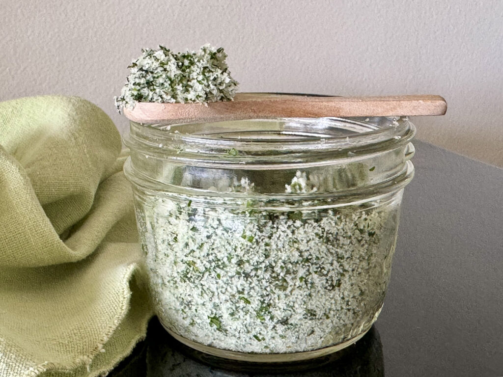 Small mason jar of filled herbed salt (salt mixed with fresh herbs) with a small wooden spoon showing a scoop.