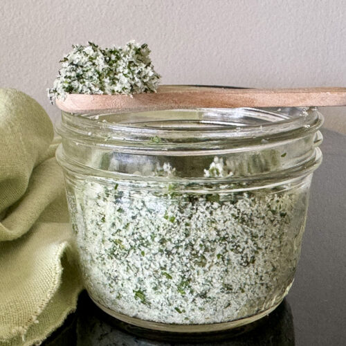 Small mason jar of filled herbed salt (salt mixed with fresh herbs) with a small wooden spoon showing a scoop.