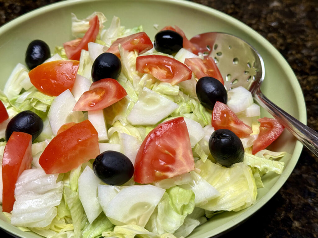 Green bowl filled with iceberg lettuce, tomatoes, cucumbers, and black olives.