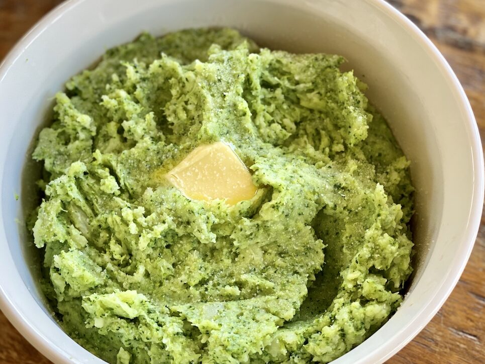A white bowl on a wood background filled with Potato-Broccoli Mash Up with a pat of melting butter.