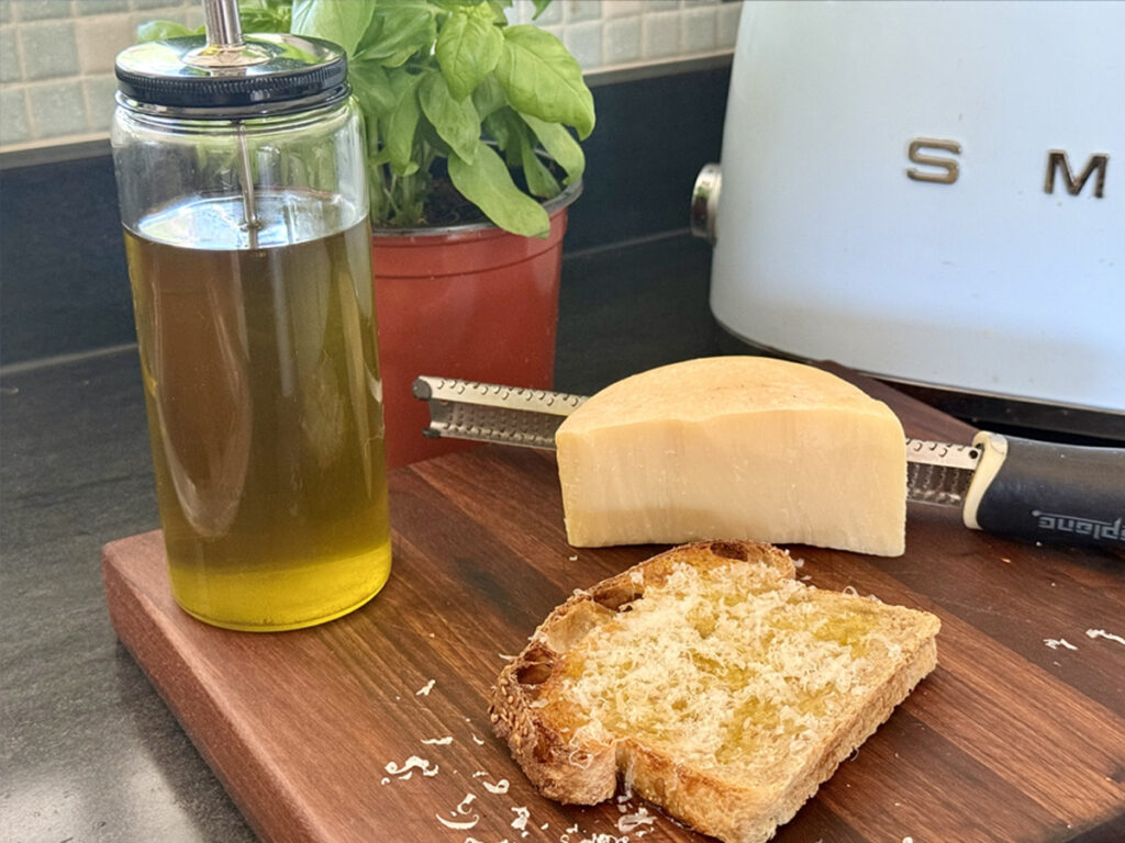 A slice of toasted sourdough drizzled with basil infused oil and topped with Grana Padano - glass bottle fill of basil oil visible.