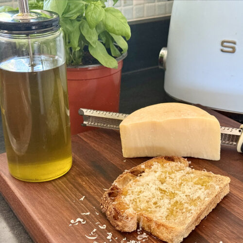 A slice of toasted sourdough drizzled with basil infused oil and topped with Grana Padano - glass bottle fill of basil oil visible.