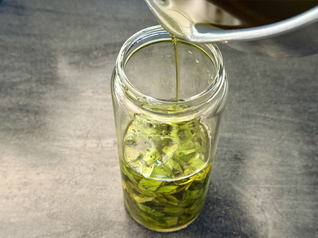 Olive oil poured over fresh chopped basil in a clear tall glass jar on a gray background.