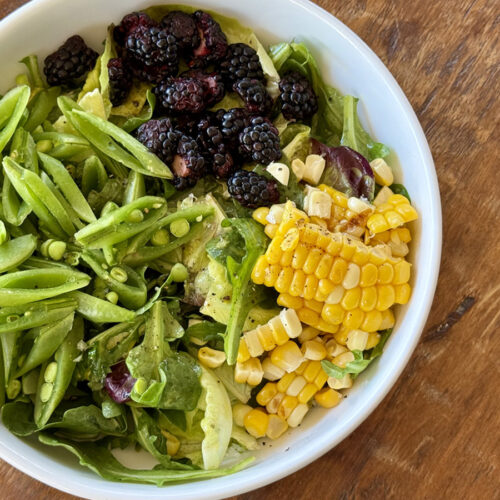 Fresh Corn off the cob, snap peas, and blackberries over mixed greens in a white bowl on a dark wood table